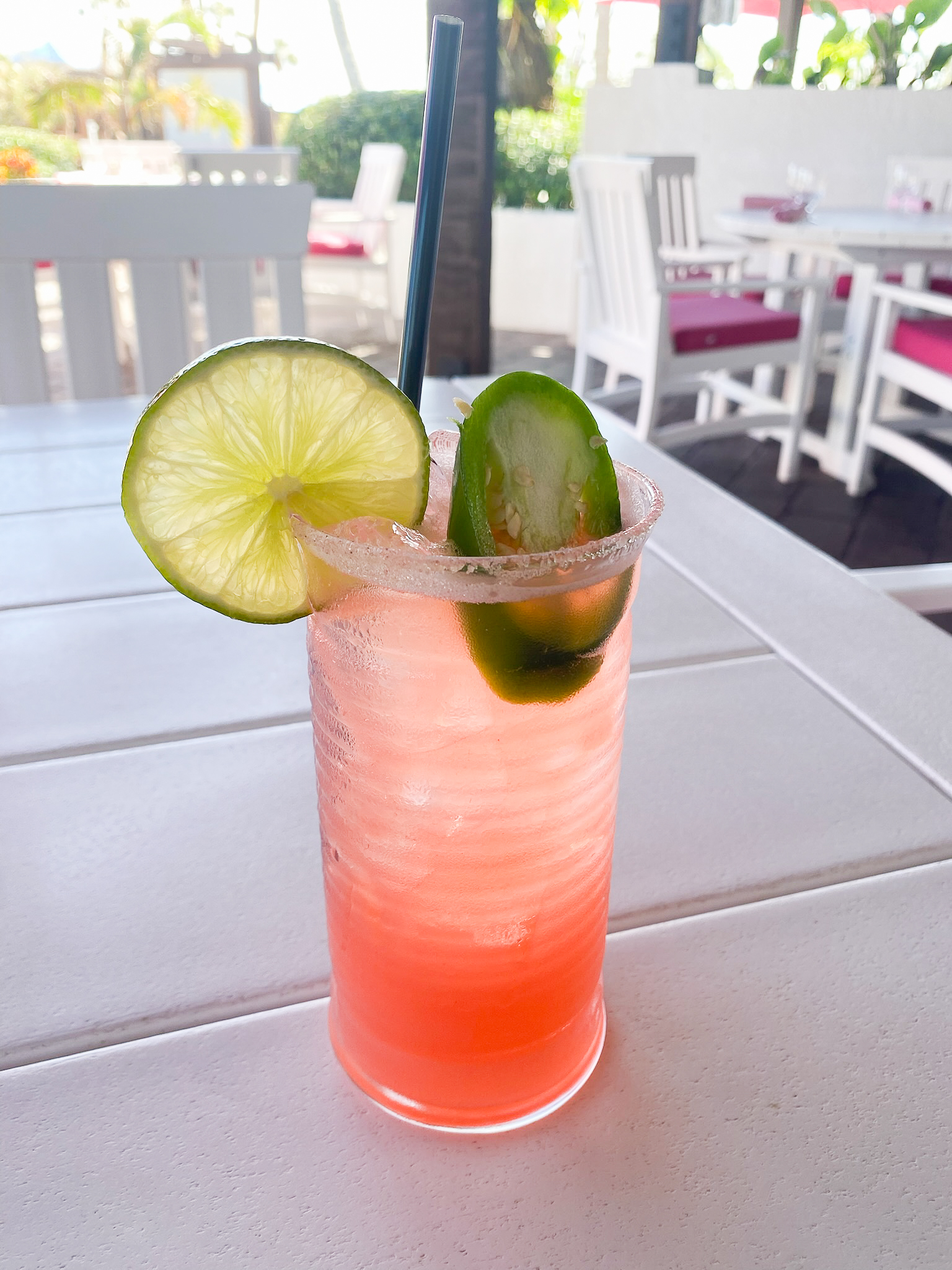 Red ombre cocktail sitting on table outdoors with a bright green lime slice and jalapeno garnish and black straw