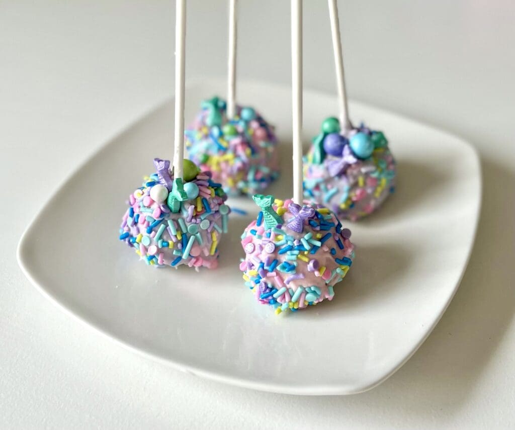 Four colorful mermaid cake pops covered in sprinkles and pink icing sitting on white plate