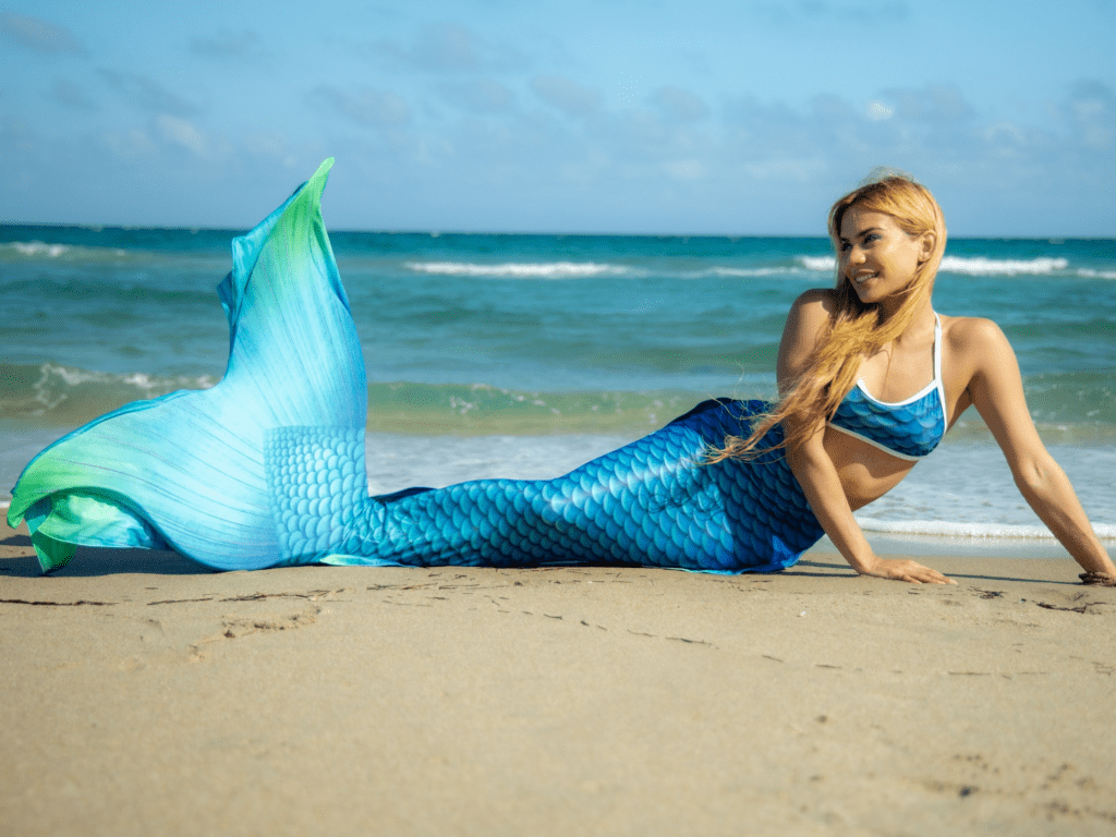 Woman in mermaid fin sitting on sandy beach