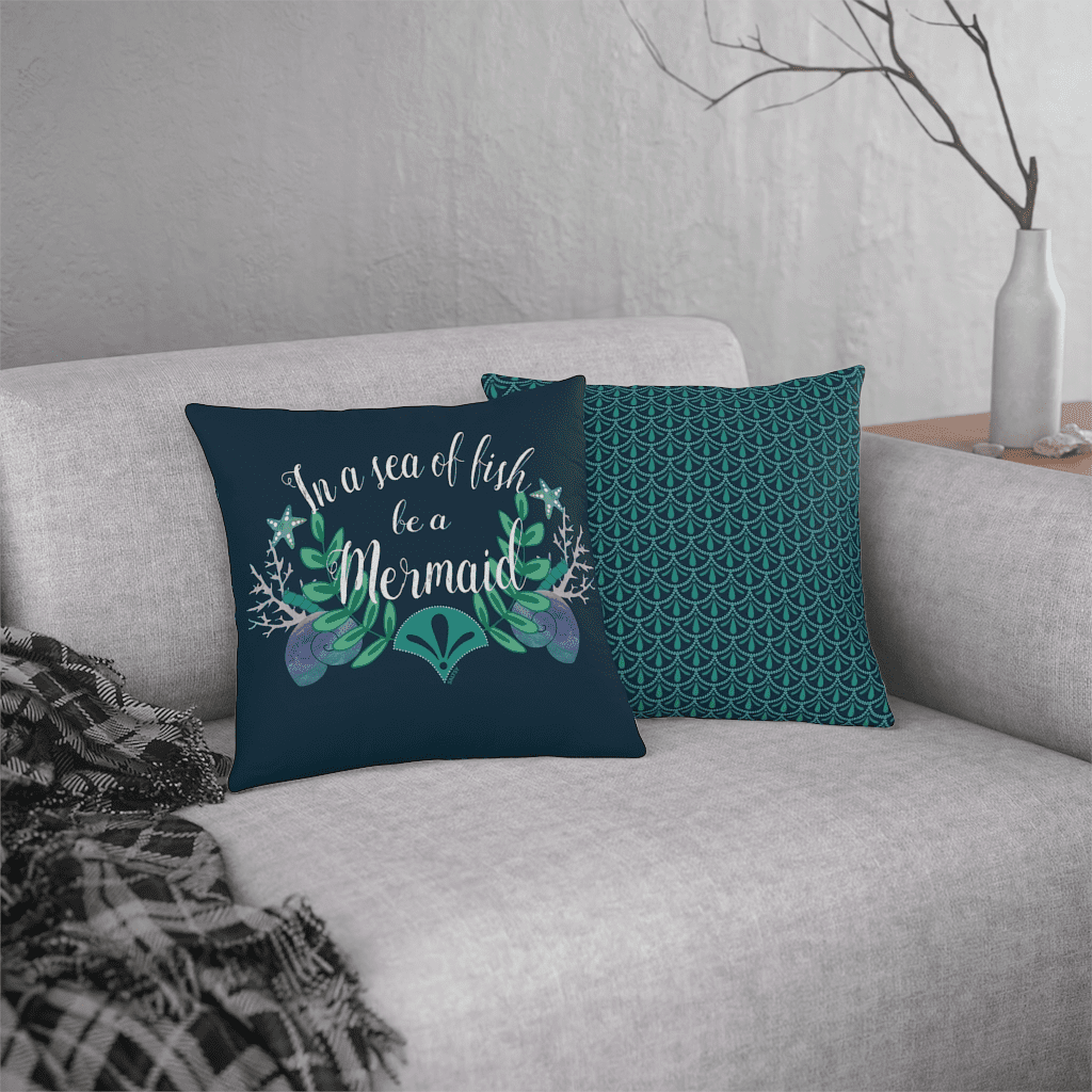 two pillows sitting on couch, one  shows the front that says "in a sea of fish, be a mermaid" with a blue background and ocean artwork, and the other is the pillow back in a dark blue and aqua mermaid print