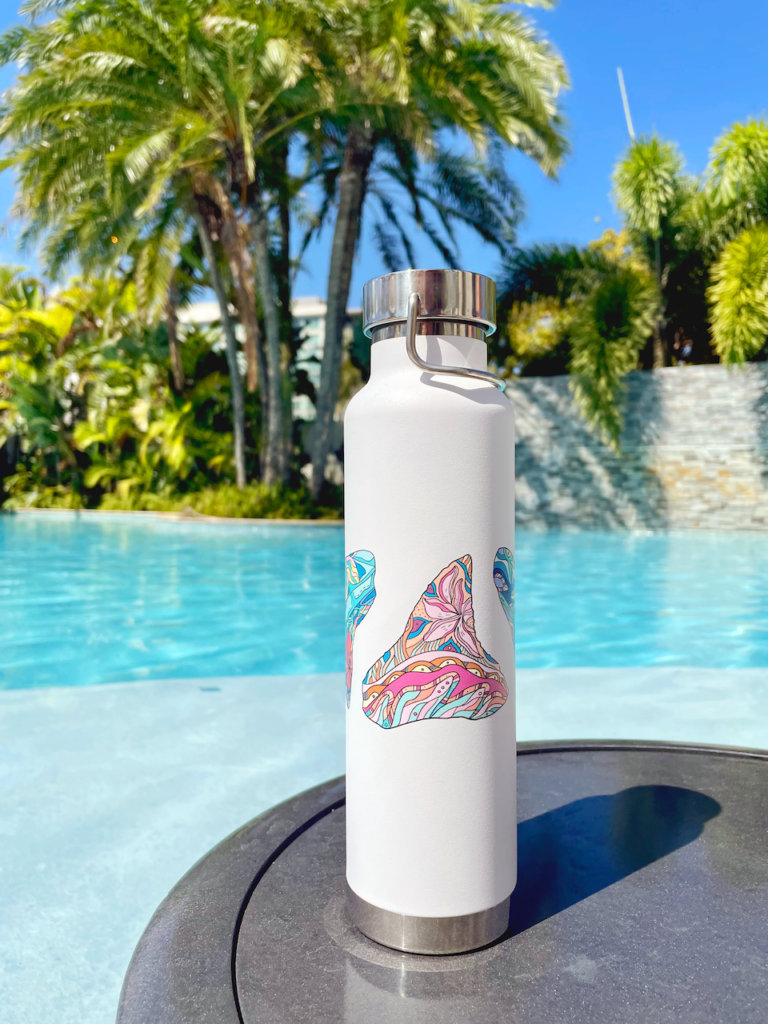 White metal water bottle with shark teeth artwork wrapping around center. Sitting on table in front of pool with palm trees.