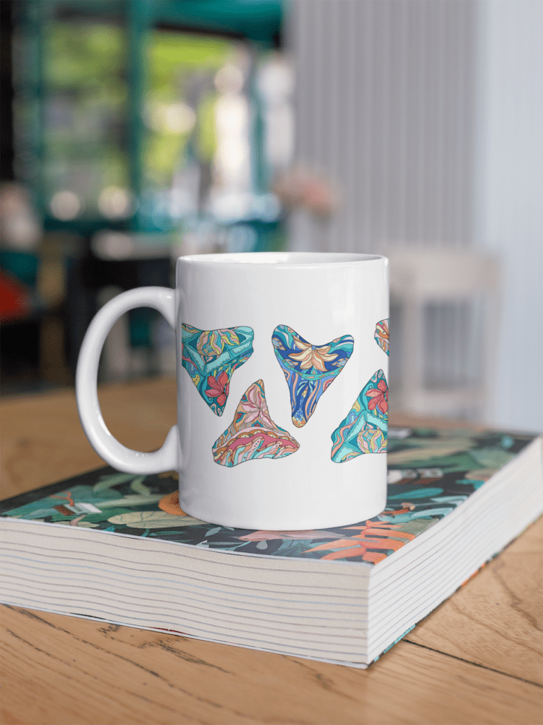 Tropical shark teeth mug sitting on book in brightly colored room. White mug with tropical shark artwork.
