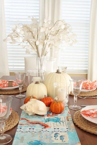 Coastal Thanksgiving tablescape with orange and white pumpkins, coral napkins, and seagrass place mats on top of an coastal themed table runner. 